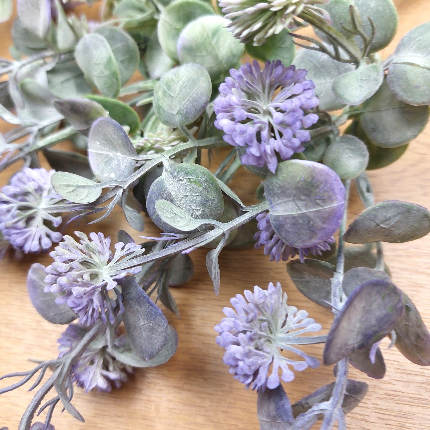Artificial floral branch with dusty purple pom-pom flowers and muted green-gray leaves on a light wood surface.