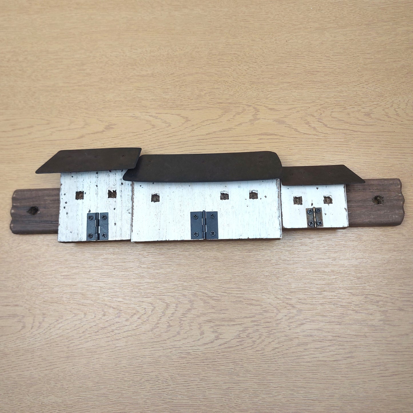 A rustic metal wall rack shaped like three croft cottages, featuring white-painted bodies, dark brown roofs, and a dark brown mounting bar, displayed on a light wood grain surface.