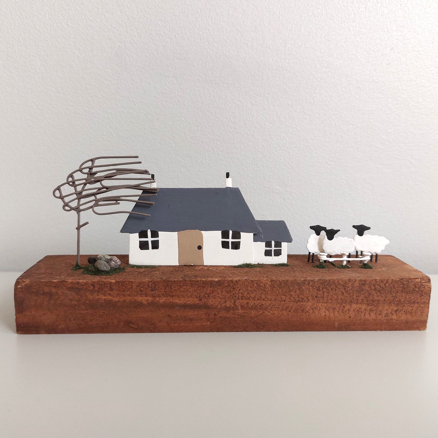 Model of a house with sheep and a tree on a wooden base against a plain background