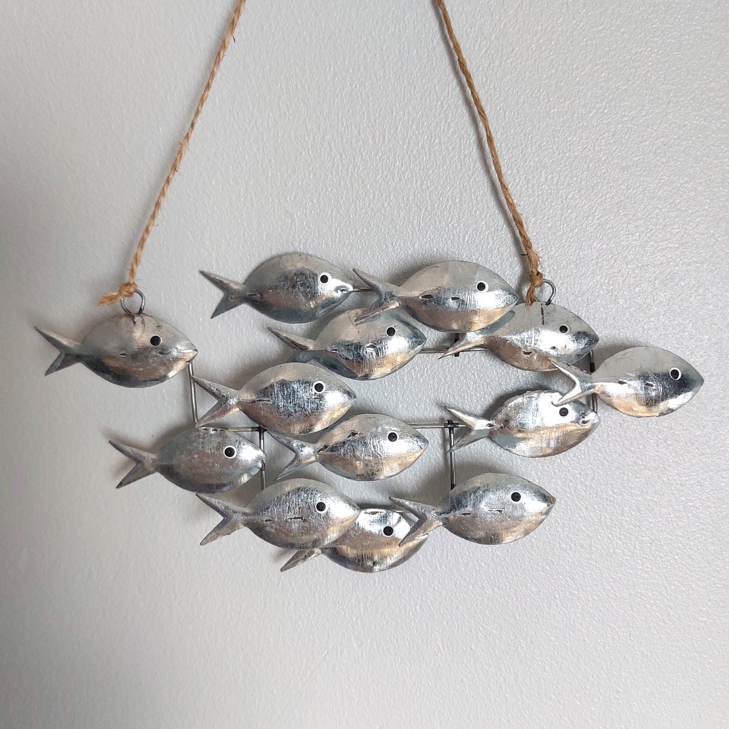 A decorative hanging wall piece made of multiple small metallic fish charms suspended from a natural twine string against a plain light wall.