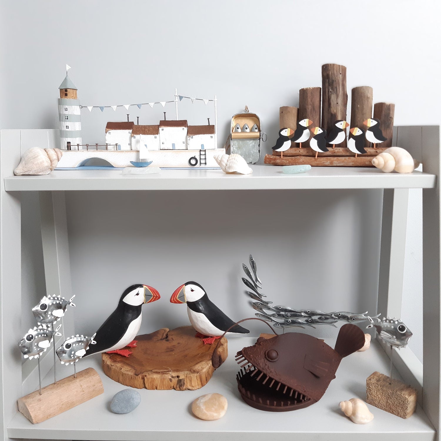 A collection of coastal decor items such as models of puffins, puffer fish and fish on light blue shelving against a pale background