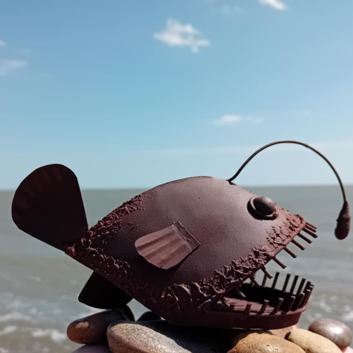 Rust Coloured Metal Angler fish Figurine on a beach with ocean and sky in the background