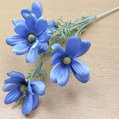 Multiple blue artificial Cosmos Daisy flowers with green centers on a light green stem, arranged on a light wood surface.