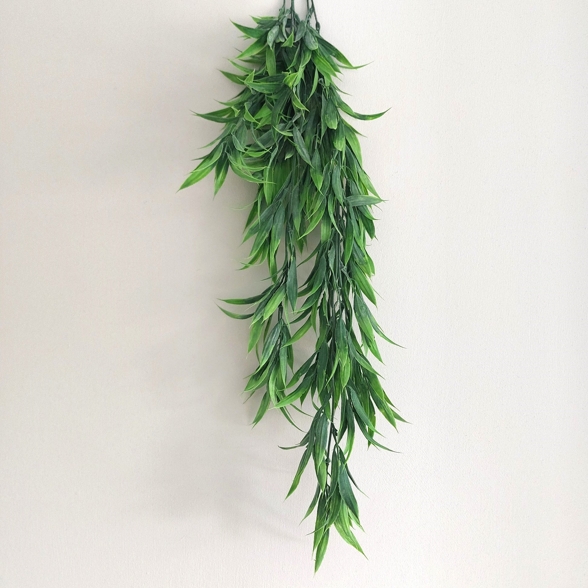 A long hanging artificial green leafy wild grass with many narrow pointed leaves, photographed against a plain light beige wall.