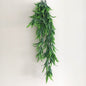 A long hanging artificial green leafy wild grass with many narrow pointed leaves, photographed against a plain light beige wall.