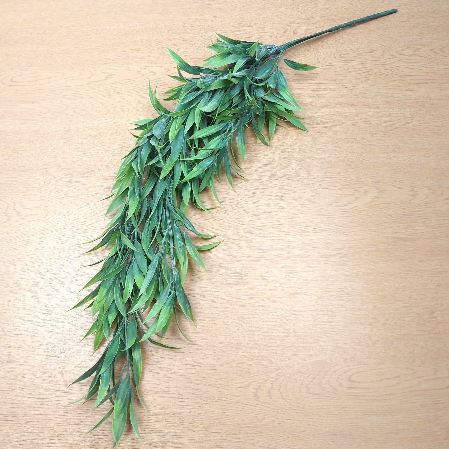 A long green leafy decorative wild grass trailing faux plant stem laid diagonally on a light wood surface.