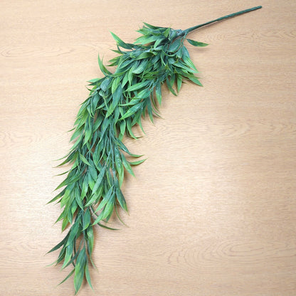 A long green leafy decorative wild grass trailing faux plant stem laid diagonally on a light wood surface.
