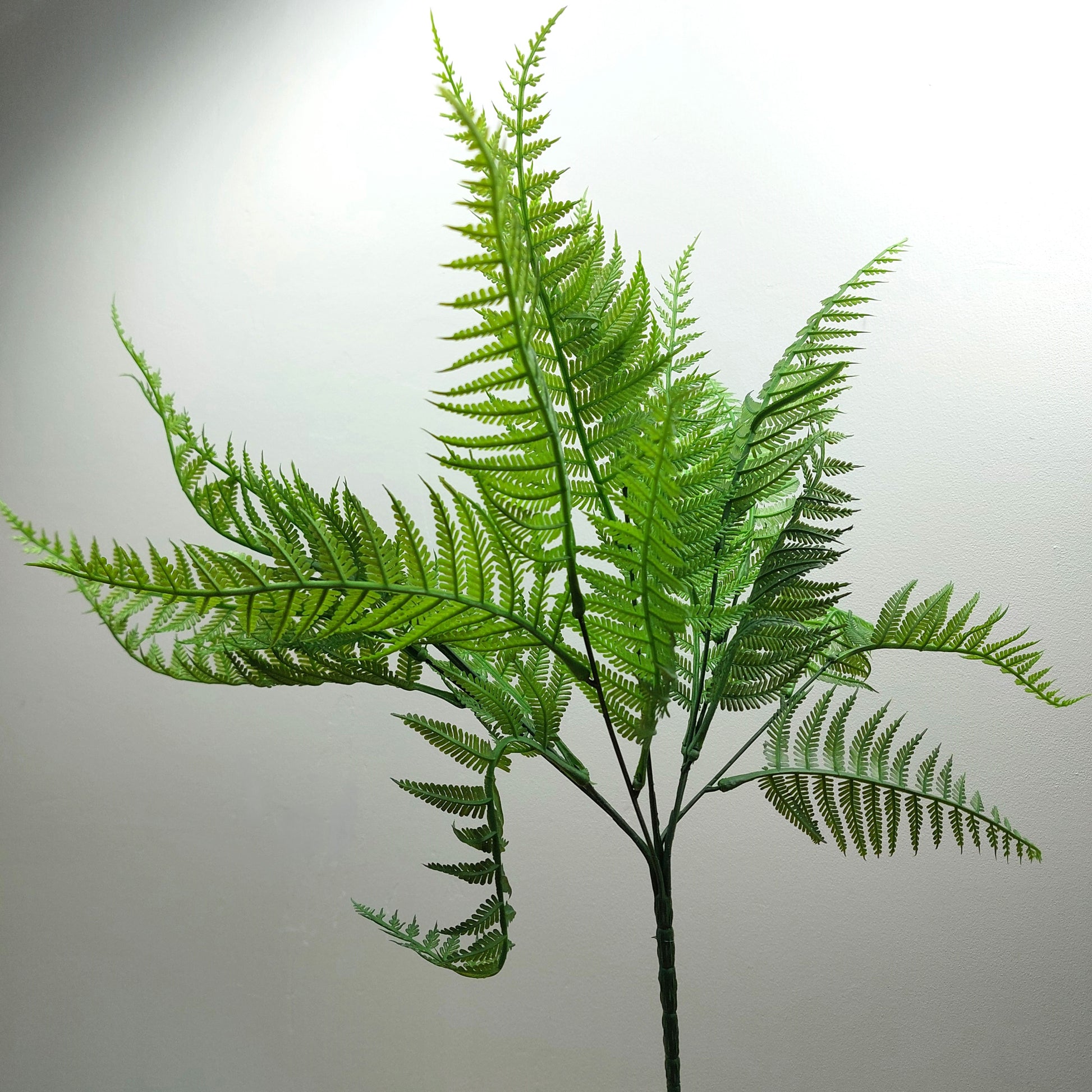 A single artificial fern stem with bright green fronds on a plain light gray background.