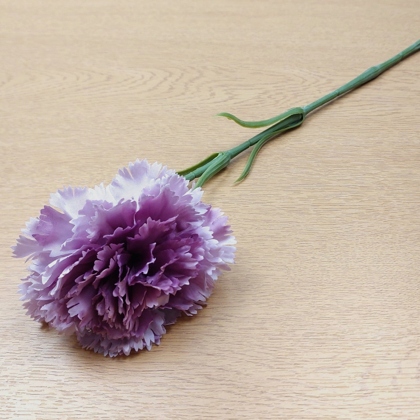 An artificial Lilac Carnation stem laying down on a wooden surface.