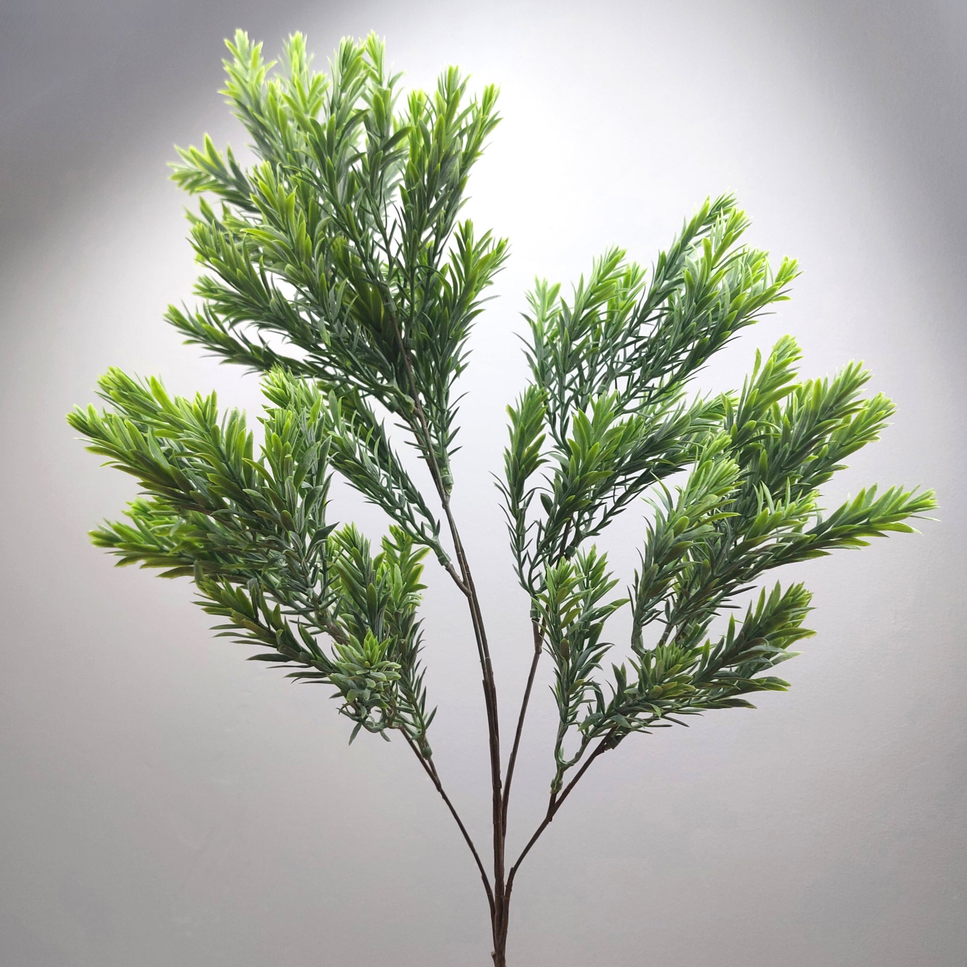 An artificial green leafy raven leaf spray or decorative plant stem with dense needle-like foliage on a plain light gray background.