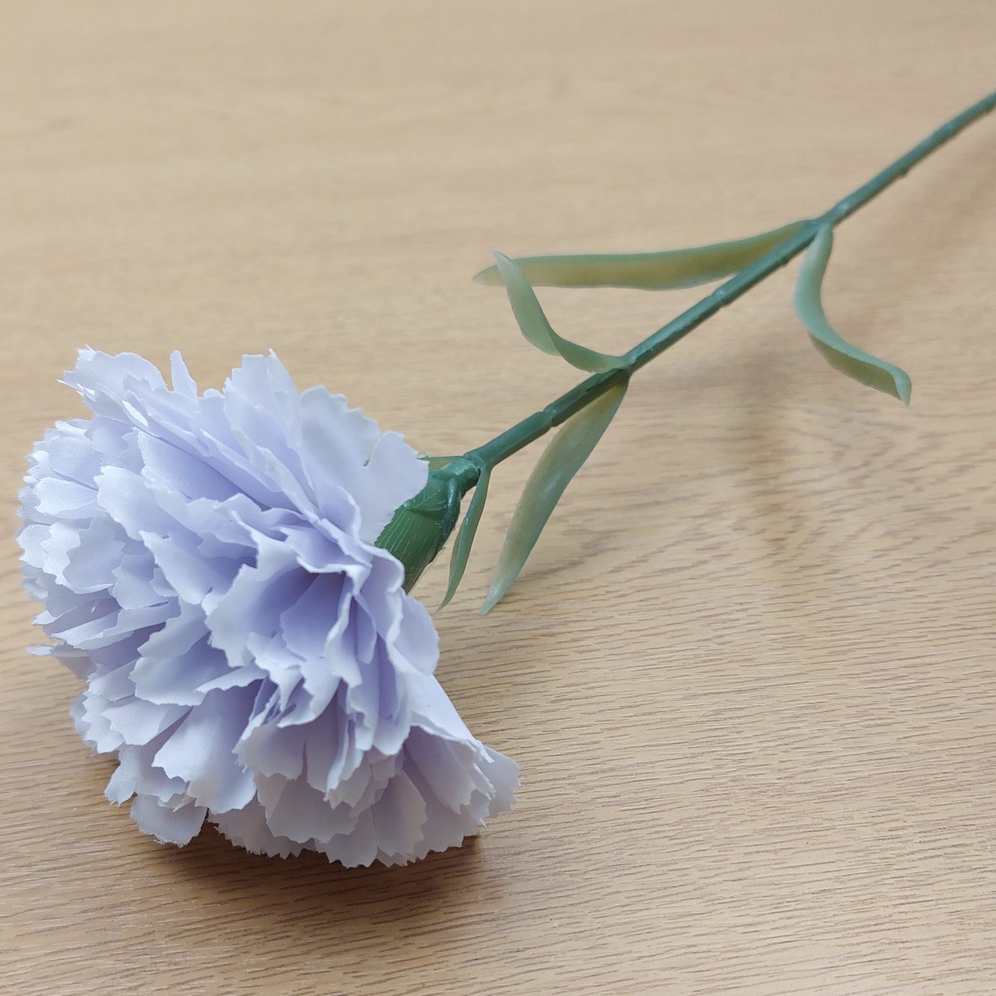 A single artificial carnation flower with pale lavender-blue petals and a green stem, placed on a light wood surface.