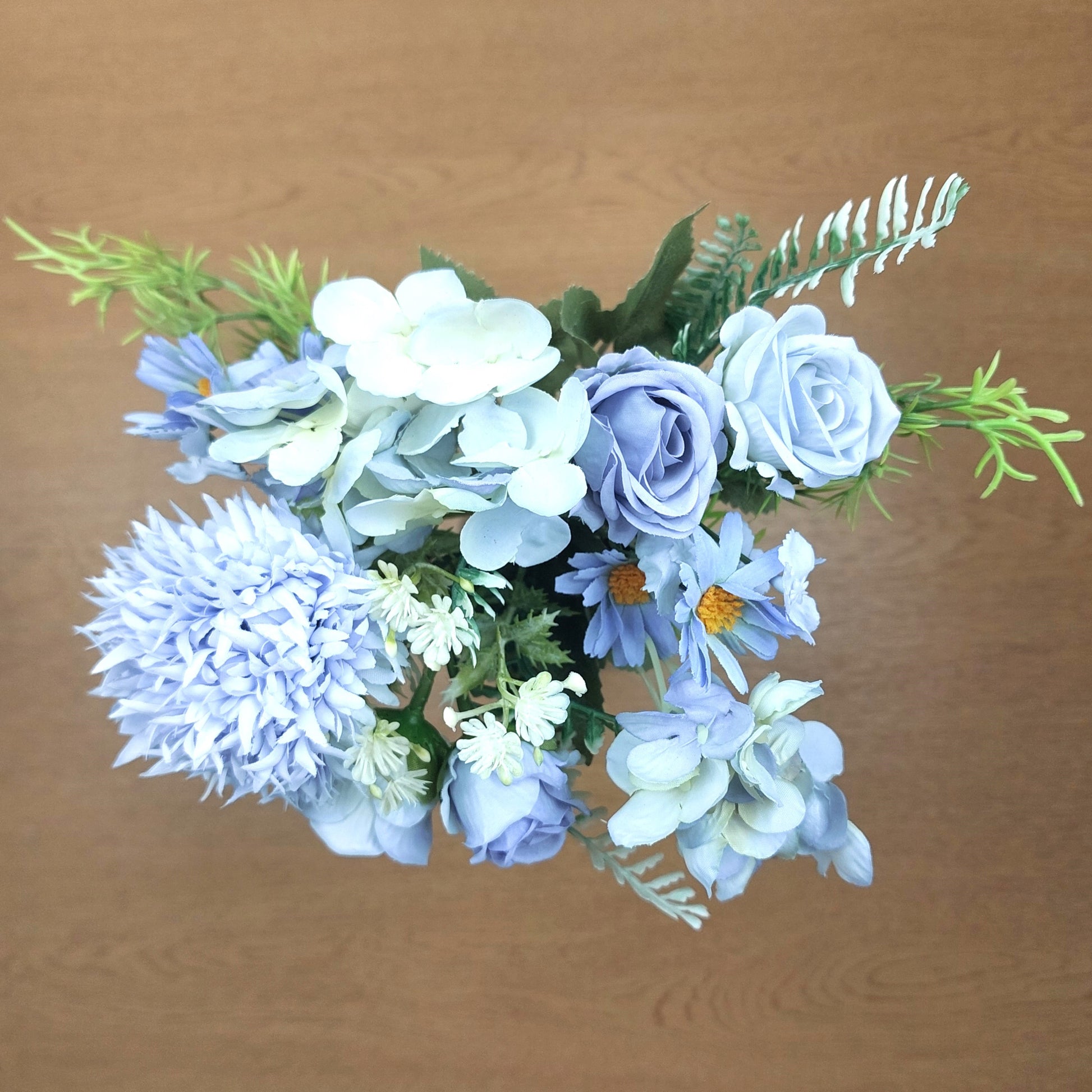 A bouquet of artificial flowers in shades of light blue and white, accented with green foliage, set against a plain brown wooden background.