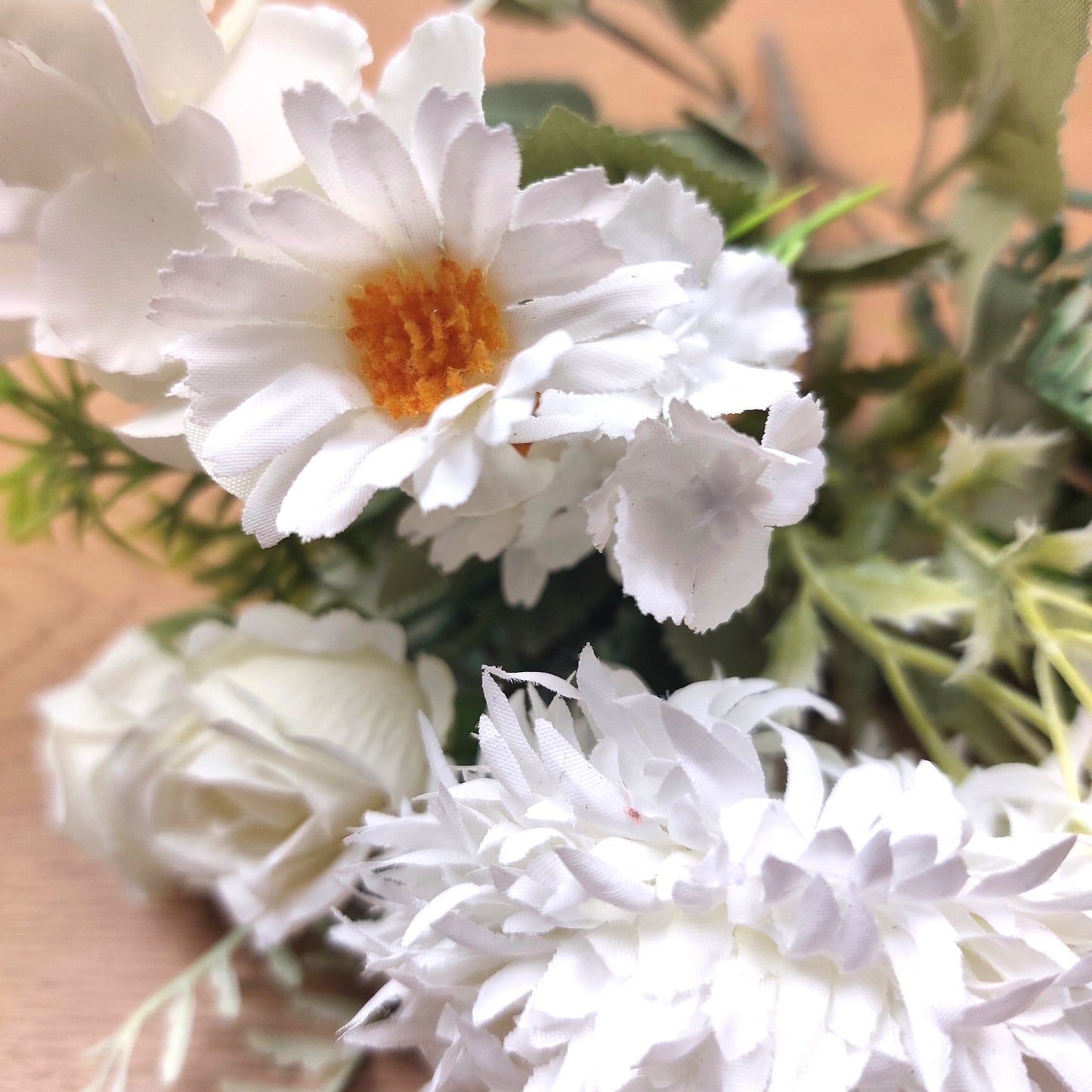 Ivory faux flower bouquet with daisy-like blooms, white roses, and green foliage on a light wood surface.