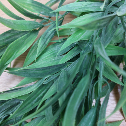 Green artificial wild grass leaves with realistic pointed tips, arranged on a light wood surface.