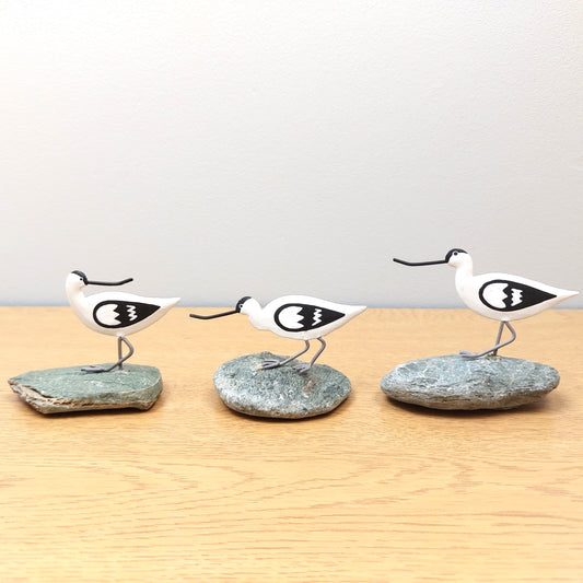 Three bird-shaped figurines on stone bases on a wooden surface with a light gray background