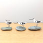 Three bird-shaped figurines on stone bases on a wooden surface with a light gray background
