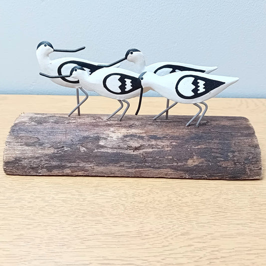 Decorative figurines of four black and white avocet birds on a wooden log against a light blue background