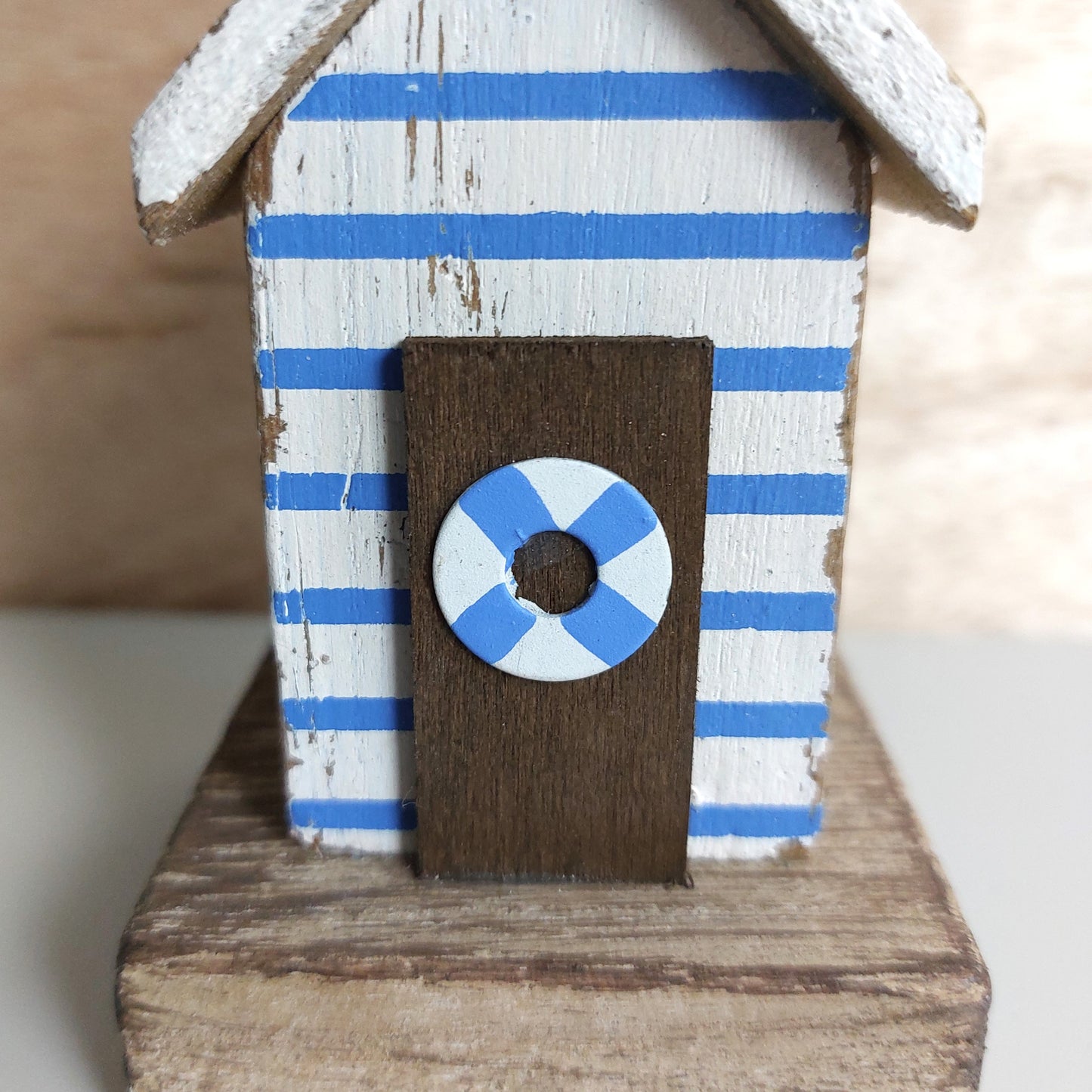 A small decorative beach house figurine with blue and white horizontal stripes, a brown door, and a blue-and-white life preserver emblem on the front, shown on a wooden base against a softly blurred neutral background.