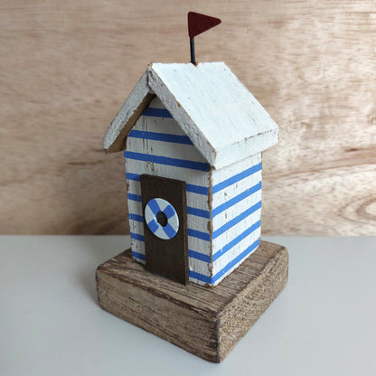 A small decorative beach hut figurine with blue and white horizontal stripes, a white roof, a dark brown door with a blue-and-white life preserver emblem, and a small dark red flag on top, displayed on a rustic wooden base against a light wood background.