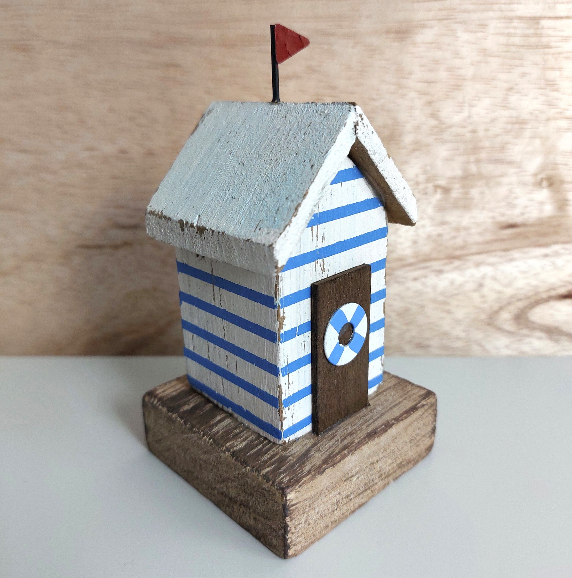 A small rustic wooden beach house figurine with blue and white horizontal stripes, a light blue-white roof, a dark brown door with a blue-and-white life preserver detail, and a tiny red flag on top, displayed against a light wood and white background.
