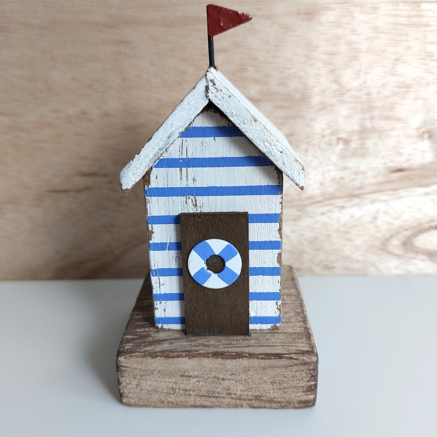 A small decorative beach hut figurine with blue and white striped sides, a white roof, a dark brown door with a blue-and-white life preserver emblem, and a small red flag on top, displayed on a wooden base against a light wood background.