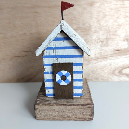 A small decorative beach hut figurine with blue and white striped sides, a white roof, a dark brown door with a blue-and-white life preserver emblem, and a small red flag on top, displayed on a wooden base against a light wood background.