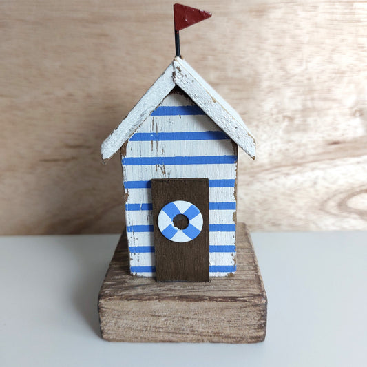 A small decorative beach hut figurine with blue and white striped sides, a white roof, a dark brown door with a blue-and-white life preserver emblem, and a small red flag on top, displayed on a wooden base against a light wood background.