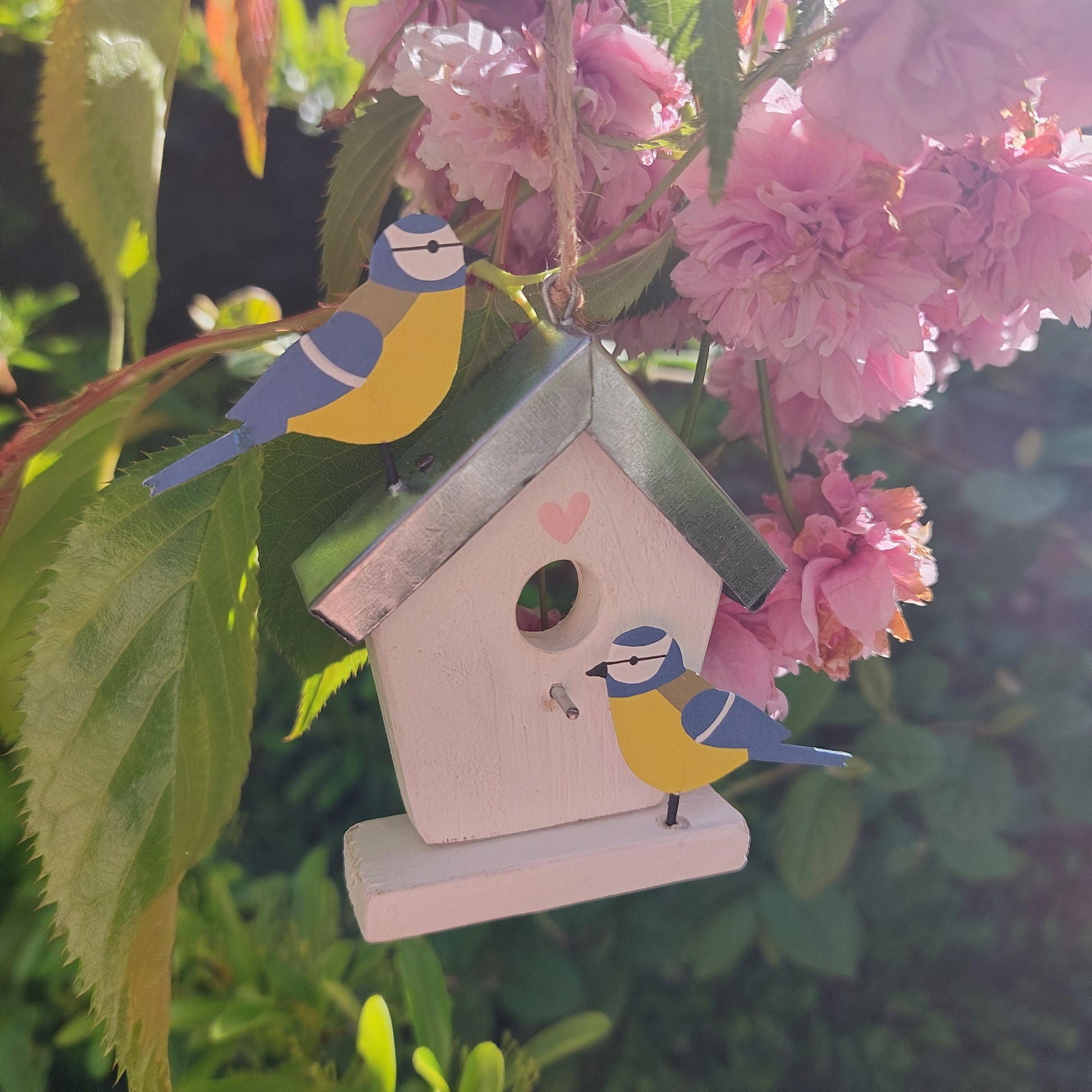 Decorative birdhouse hanging with metal birds on a branch with pink flowers and green leaves.
