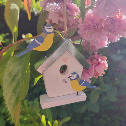 Decorative birdhouse hanging with metal birds on a branch with pink flowers and green leaves.