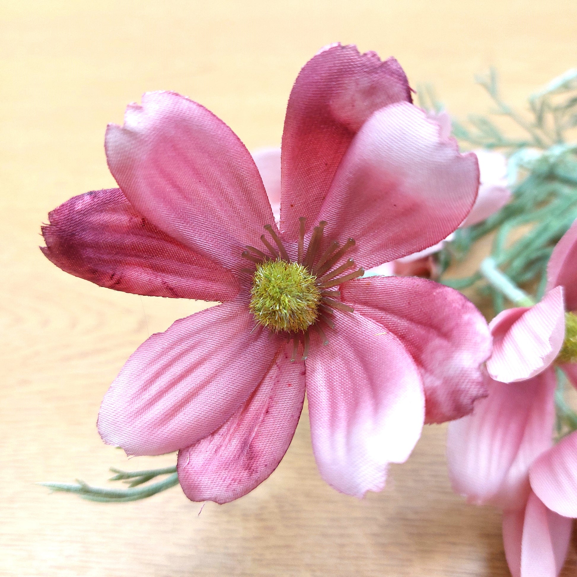 A close-up of a pink silk fake cosmos flower with layered petals and a green-yellow center, shown on a light wood surface with other similar flowers blurred in the background.