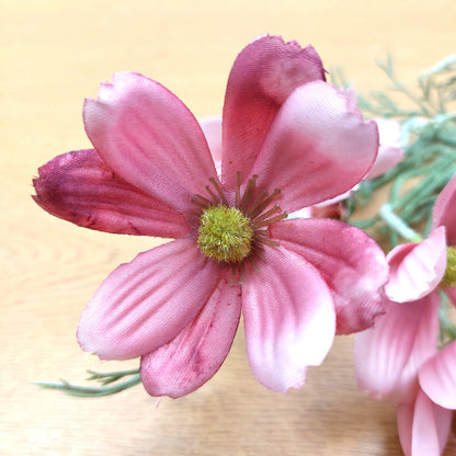 A close-up of a pink silk fake cosmos flower with layered petals and a green-yellow center, shown on a light wood surface with other similar flowers blurred in the background.