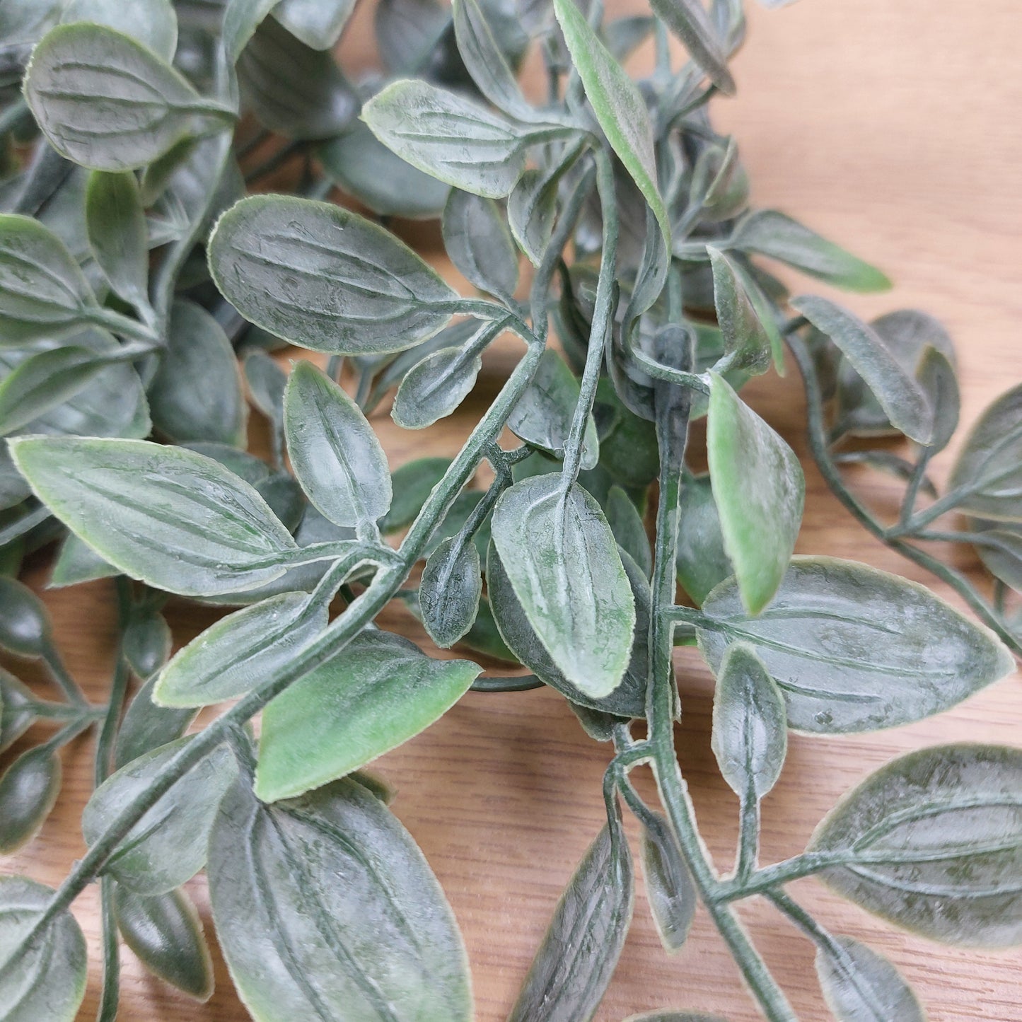 Artificial green leafy ruscus stem with textured, dusty gray-green leaves on a light wood background.