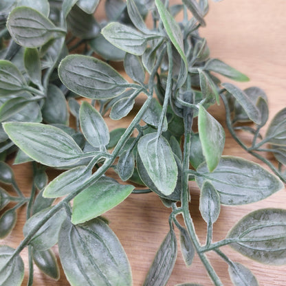 Artificial green leafy ruscus stem with textured, dusty gray-green leaves on a light wood background.