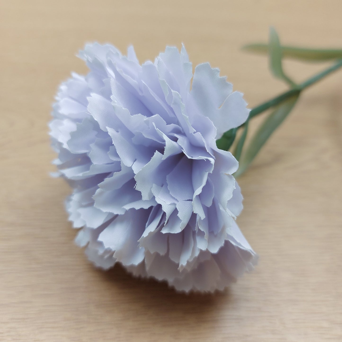 A close up of an artificial single pale lavender carnation flower with layered ruffled petals and a green stem, photographed on a light wood surface.