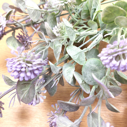 Faux artificial flower branch with dusty purple flower clusters and muted green-gray leaves on a light wood surface.