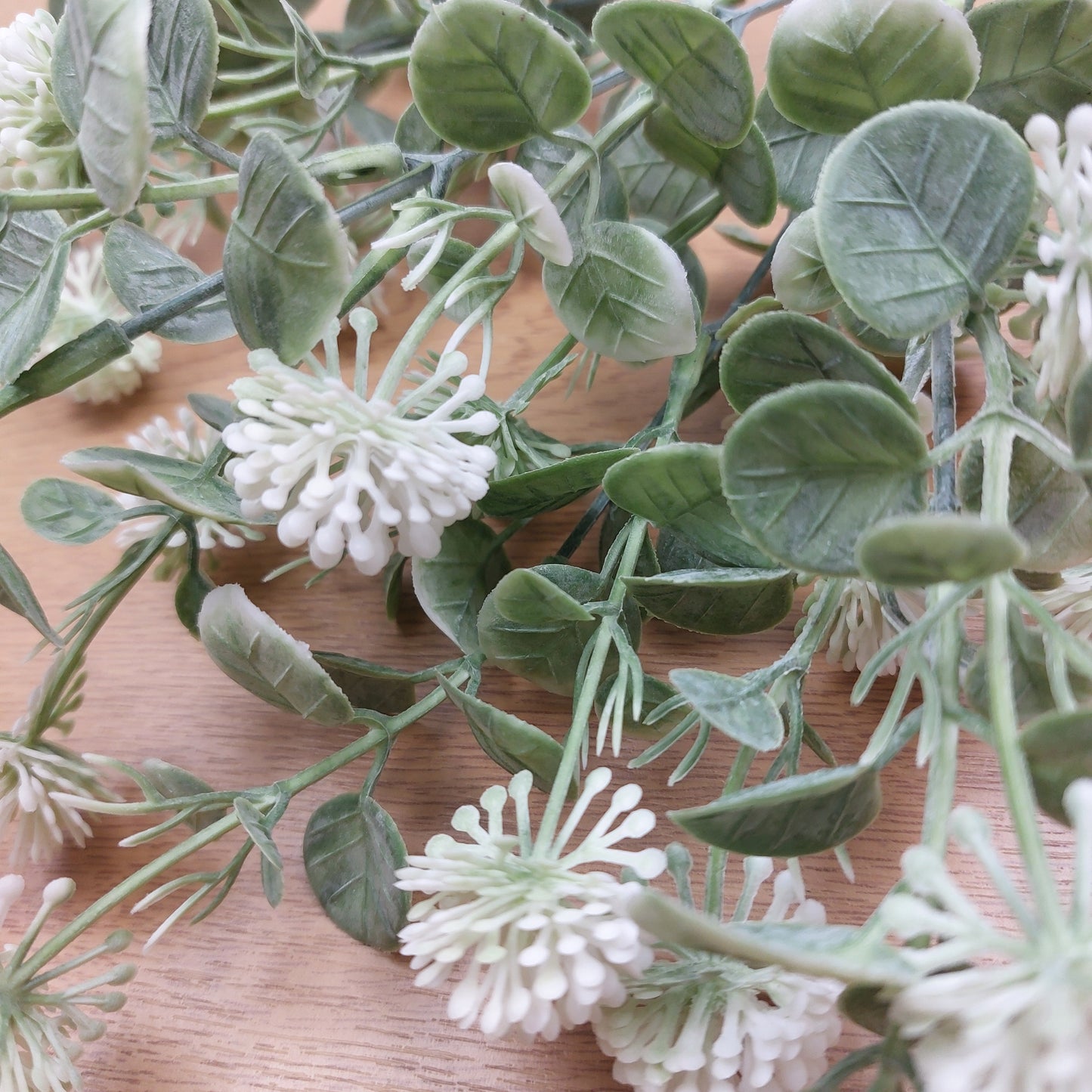 Artificial green and white floral spray with oval green leaves and clusters of small white and green flowers, arranged on a light wood surface.