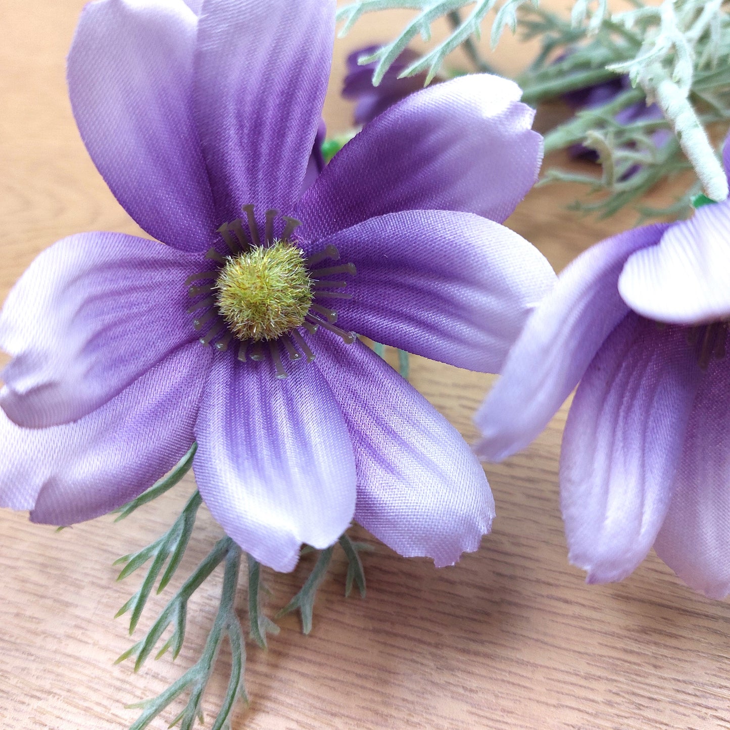 Purple Lilac artificial Cosmos flowers with pale lavender edges and a green-yellow center, attached to green stems, photographed close up on a light wood surface.