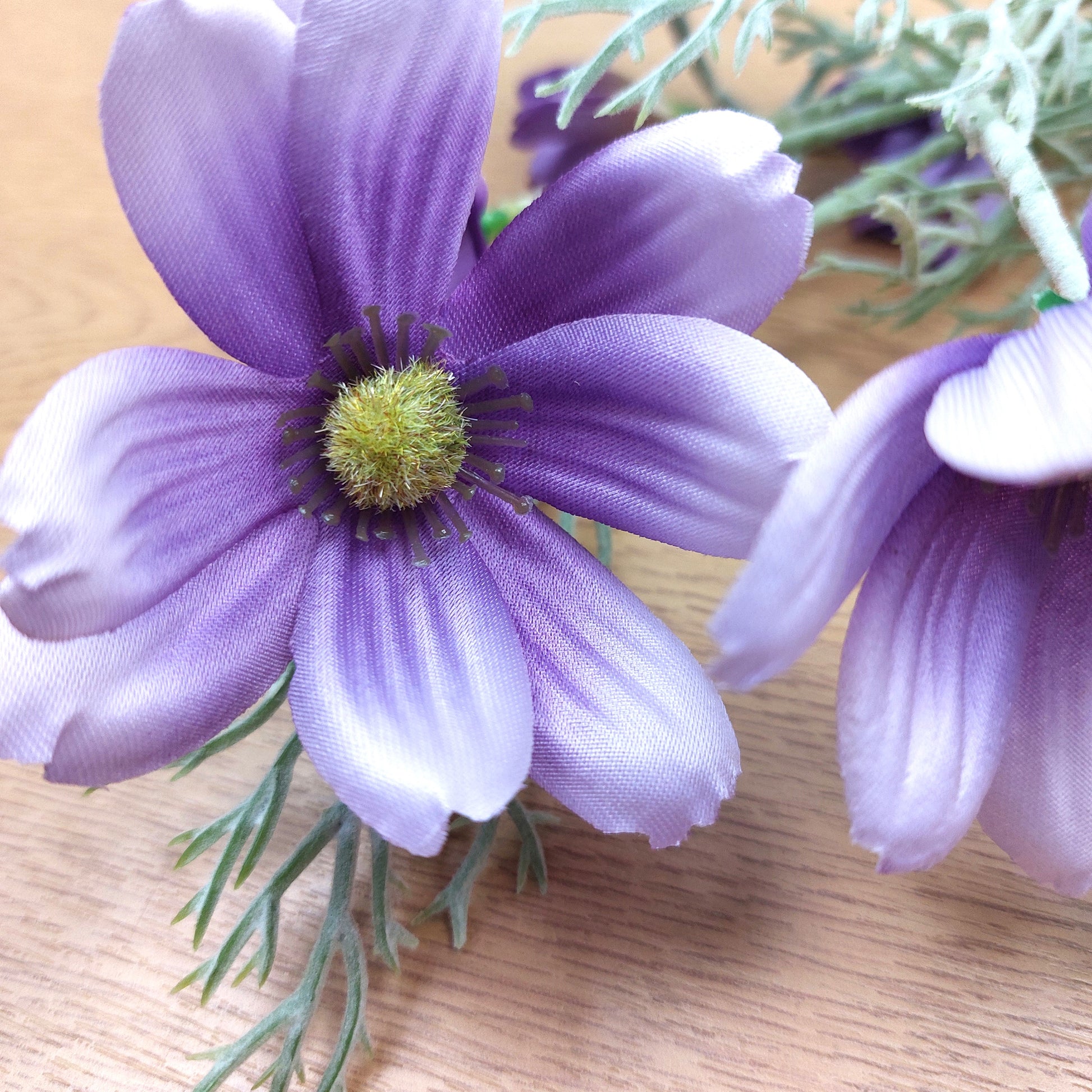 Purple Lilac artificial Cosmos flowers with pale lavender edges and a green-yellow center, attached to green stems, photographed close up on a light wood surface.