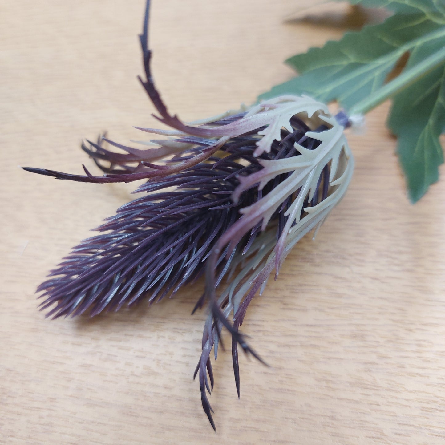 A close-up of a faux purple eryngium sea holly head with feathery fronds, placed on a light wood surface.