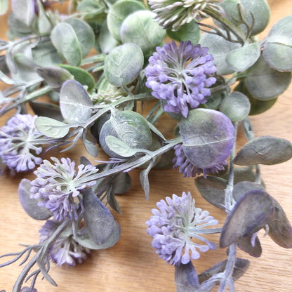 Artificial floral branch with dusty purple pom-pom flowers and muted green-gray leaves on a light wood surface.