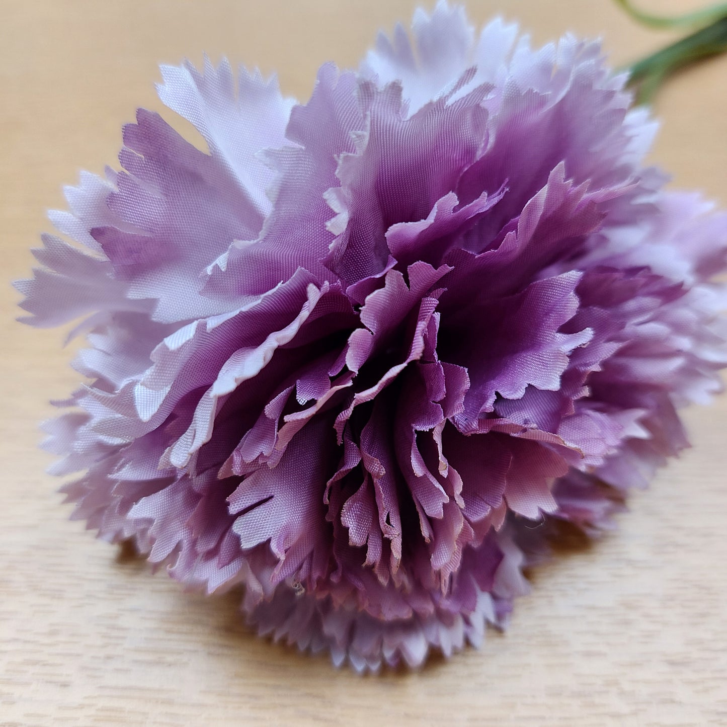 A close-up of a faux ruffled carnation flower with layered petals in shades of purple, lilac and white, photographed on a light wood surface.