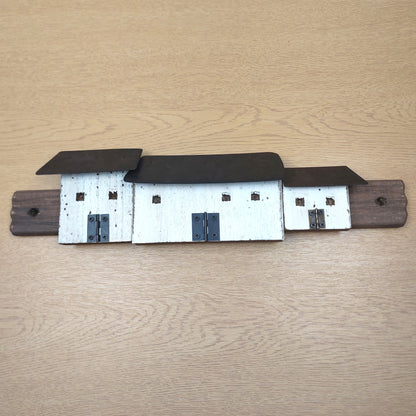 A rustic metal wall rack shaped like three croft cottages, featuring white-painted bodies, dark brown roofs, and a dark brown mounting bar, displayed on a light wood grain surface.