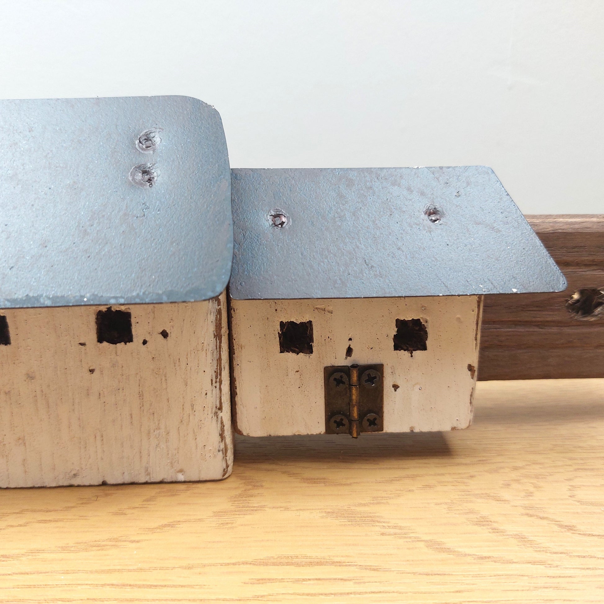 A close-up of a miniature wooden house model with a weathered light blue-gray roof and off-white painted sides, resting on a light wood surface against a plain light background.