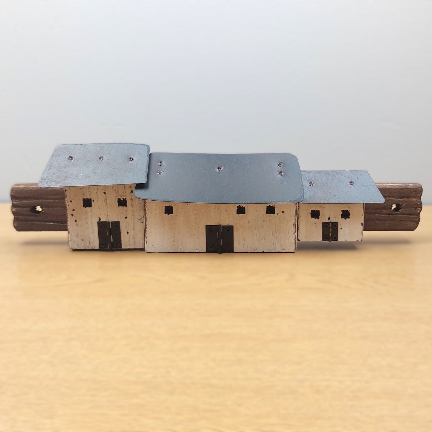 A miniature wooden model of a set of croft cottages with a gray metal roof and light wood walls, displayed on a light wooden surface against a neutral gray background.