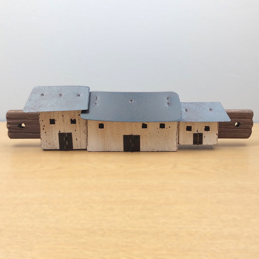 A miniature wooden model of a set of croft cottages with a gray metal roof and light wood walls, displayed on a light wooden surface against a neutral gray background.