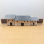 A miniature wooden model of a set of croft cottages with a gray metal roof and light wood walls, displayed on a light wooden surface against a neutral gray background.