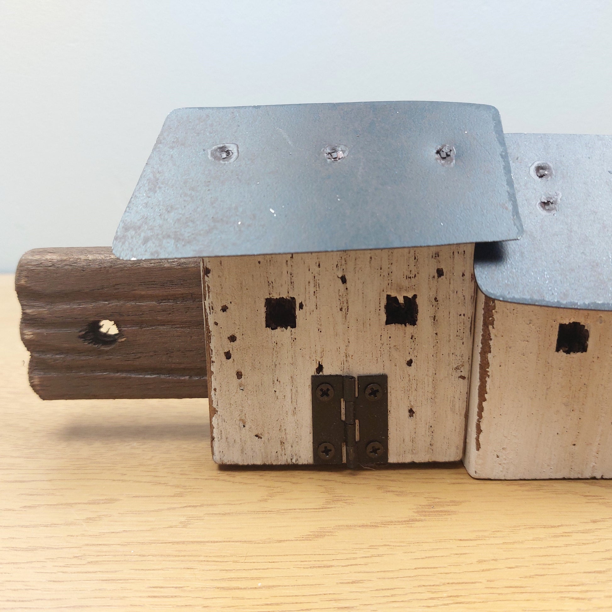 A rustic wooden house figurine with a weathered beige-painted front, dark brown side wall, and a galvanized metal roof, displayed on a light wood surface against a neutral background.
