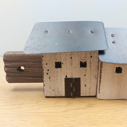 A rustic wooden house figurine with a weathered beige-painted front, dark brown side wall, and a galvanized metal roof, displayed on a light wood surface against a neutral background.