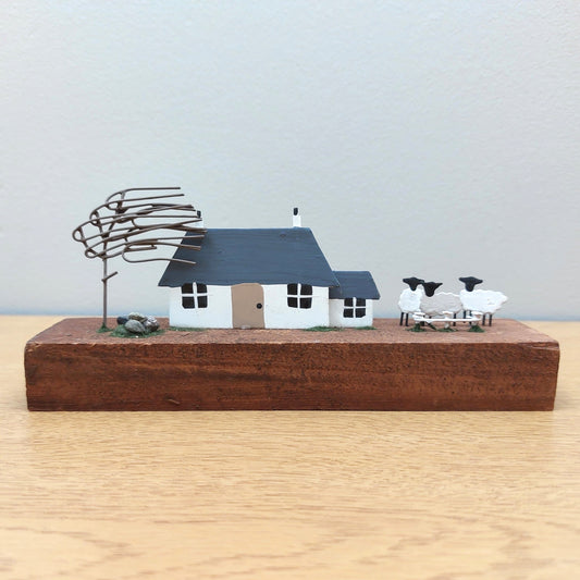 Ornamental wooden and metal scene of a windswept croft with a farmhouse, tree, sheep and stones on a wooden block against a grey background