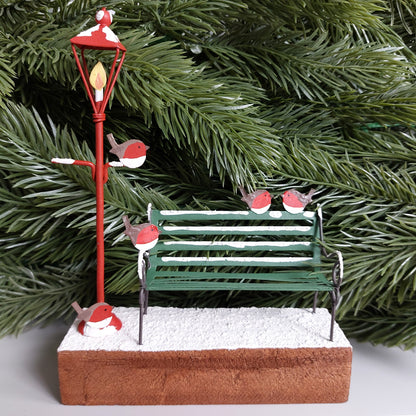 Decorative Christmas scene with a red lamp post, green bench, and red robins on a snowy wooden base against a faux green spruce tree background.